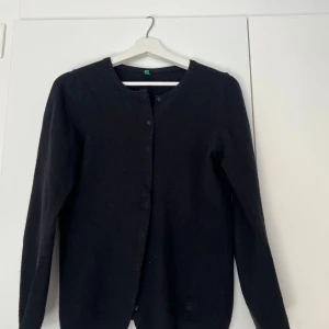 Svart kofta från Benetton i ull - Säljer en svart kofta från United Colors of Benetton! Materialet är av mjuk ull. Koftan har rund halsringning, knäppning framtill med diskreta knappar och långa ärmar. Den är använd, men fortfarande i bra skick!