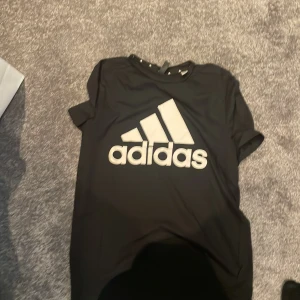 Svart Adidas t-shirt med stor logga - Svart t-shirt från Adidas med stor vit logga på bröstet. Klassisk rund hals och korta ärmar. Tillverkad i mjuk polyester som andas, perfekt för träning eller chill. Diskret 'AEROREADY'-tryck på ryggen. Snygg och sportig stil för dig som gillar streetwear.