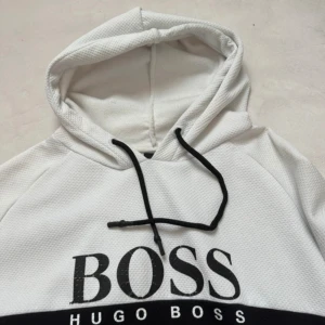 Vit och svart hoodie från Hugo Boss - Snygg hoodie från Hugo Boss i vit och svart färgblock-design. Tröjan har stor BOSS-logga framtill, huva med svarta snören och är tillverkad i ett strukturerat, sportigt material. Perfekt för dig som gillar streetwear och vill sticka ut med en clean look.