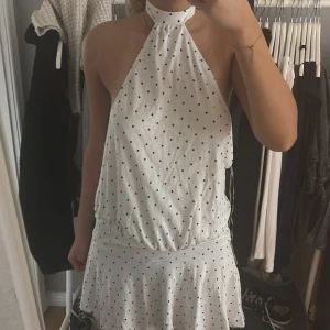 Vit prickig halterneckklänning - Säljer en vit kortklänning med svarta prickar och halterneck. Klänningen har öppen rygg, knyt i nacken och volangkant nertill. Sval och luftig modell som sitter löst över kroppen. Perfekt för varma dagar och somriga tillfällen.