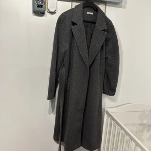 Mörkgrå lång kappa med bälte - Snygg mörkgrå kappa från NA-KD med klassisk krage och bälte i midjan. Lång modell med raka linjer och diskreta fickor. Perfekt för dig som gillar en stilren och tidlös look. Fodrad insida för extra komfort och värme.