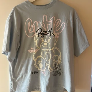 Ljusblå t-shirt med björntryck H&M - Ljusblå t-shirt från H&M med stort tryck av en tecknad björn och text i rosa och svart framtill. Oversized passform och korta ärmar. Tillverkad i 100% bomull, perfekt för en chill och trendig look.