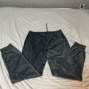 Svarta mjukisbyxor från EA7 - Säljer ett par mörk grå  mjukisbyxor från EA7 med elastisk midja och snörning. Byxorna har ribbade muddar vid bensluten och praktiska fickor. Materialet är mjukt och skönt, perfekt för chill eller träning. EA7-logga på ena benet för en clean look. Pris kan diskuteras vid snabb affär