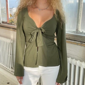 Olivgrön långärmad knytblus - Trendig olivgrön blus med djup v-ringning och snygg knytning framtill. Långa ärmar och lätt utsvängd passform ger en avslappnad men chic vibe. Perfekt att styla med jeans eller kjol för en cool look. PRIS GÅR ATT DISKUTERA 