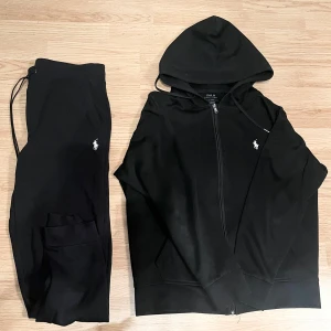 Svarta mjukisbyxor och hoodie från Polo Ralph Lauren - Svarta mjukisbyxor från Polo Ralph Lauren med vit broderad logga på benet. Byxorna har dragsko i midjan och praktiska fickor på sidorna. Perfekta för chill eller träning, mjukt material och stilren look.