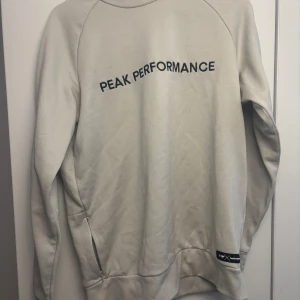 Beige Peak Performance sweatshirt - Snygg beige sweatshirt från Peak Performance med svart logotyp tryckt över bröstet. Tröjan har långa ärmar, rund hals och diskret dragkedja vid sidan. Perfekt för chill eller träning, tillverkad i mjuk bomullsmix för skön komfort.