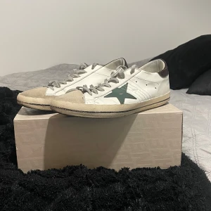 Golden Goose sneakers med grön stjärna - Golden Goose sneakers i vitt skinn med grå snörning och beige mockadetaljer på tån. Sidorna har en grön stjärna och hälen är mörkbrun med textur. Smått slitage på sulan, annars väldigt bra skick ⭐️Köpta på Merch Sweden förra sommaren, har en orderbekräftelse