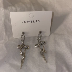 Silvriga örhängen med rosett och spike - Snygga silvriga örhängen med hängande rosett och spetsig spike-detalj. De har en cool kedja mellan pluppen och rosetten, vilket ger en edgy vibe. Perfekta för dig som vill sticka ut med accessoarerna.