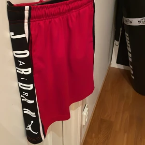 Röda Jordan shorts med logga - Snygga röda Jordan shorts med bred svart sidopanel och vit logga längs benet. Elastisk midja med snörning för skön passform. Tillverkade i mjuk polyester som andas, perfekta för sport eller chill. Klassisk Jumpman-detalj på sidan ger extra streetkänsla. Är använda några gånger men i bra skick.