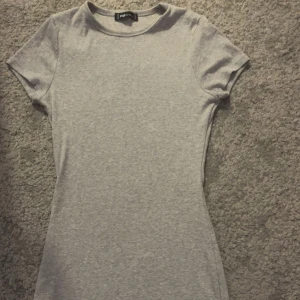 Grå kortärmad klänning från F21 - En enkel och stilren grå klänning från F21. Modellen är kort, har rund halsringning och korta ärmar. Perfekt för dig som gillar en clean och avslappnad look. Materialet är mjukt och stretchigt, vilket gör den bekväm att bära hela dagen. Storlek M men väldigt liten i storlek funkar till S och XS