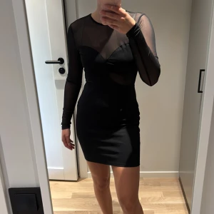 Svart meshklänning från NA-KD - Supersnygg svart kort klänning från NA-KD med meshdetaljer upptill och på ärmarna. Långärmad och figurnära modell med coola transparenta partier som ger en edgy vibe. Perfekt för dig som vill sticka ut med en modern look.