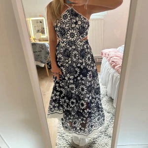 Blommig långklänning - Supersnygg långklänning med svart och vitt blommönster, halterneck och fina cut-outs i midjan. Klänningen har en flowy kjol och är ärmlös, perfekt för sommarens alla tillfällen. Materialet känns lätt och luftigt.