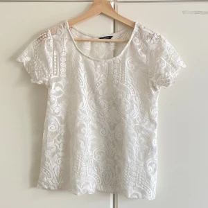 Vit spetsblus från Lindex - Säljer en superfin vit blus från Lindex med korta ärmar och rund halsringning. Blusen är helt täckt av vacker spets med detaljerat mönster och har en luftig känsla, perfekt för varma dagar eller när du vill känna dig extra söt.
