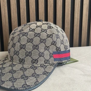 Gucci beige keps med GG-mönster - Snygg keps från Gucci i beige och mörkblått med klassiskt GG-monogram över hela kepsen. På sidorna finns Guccis ikoniska röd-blå rand. Kepsen har böjd skärm och justerbar rem baktill. Materialet är bomull och polyester, insidan är mörkblå.