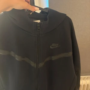 Svart Nike jacka med dragkedja XL 158-170cm - Svart jacka från Nike i storlek XL (Nike tech), med hel dragkedja framtill och diskret Nike-logga på bröstet. Jackan har en sportig look med en vågig detalj över bröstet och hög krage. Perfekt för dig som gillar streetwear och enkel stil.