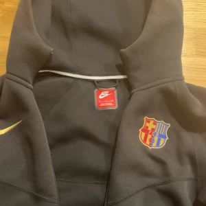 Svart Nike FC Barcelona hoodie - Svart hoodie från Nike med FC Barcelona-logga på bröstet och gul Nike swoosh. Dragkedja framtill, två fickor med dragkedja och huva. Spotify-logga på ena ärmen. Perfekt för dig som älskar fotboll och vill ha en sportig look.