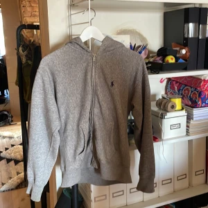 Grå hoodie från Polo Ralph Lauren - Grå hoodie från Polo Ralph Lauren med dragkedja framtill och klassisk huva. Tröjan har långa ärmar, ribbade muddar och den ikoniska blå Polo-loggan broderad på bröstet. Perfekt för chill dagar och enkel att matcha med jeans eller joggers.