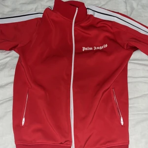 Röd vindjacka från Palm Angels M - Palm Angels röd vindjacka med vita detaljer och dragkedja framtill. Jackan har vita ränder längs ärmarna och logga på bröstet. Tillverkad i bomull och polyester, med två fickor med dragkedja. Perfekt för en sportig och trendig look.