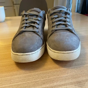 Grå mockasneakers från Christian Louboutin - Snygga grå sneakers i mocka från Christian Louboutin med klassisk röd sula och vita kanter. Skorna har rund tå, snörning och diskreta detaljer. Perfekta för dig som vill ha en stilren och exklusiv look med en touch av lyx. Dem är 24,5 cm Nypris ca 8000. Mitt pris 1600kr