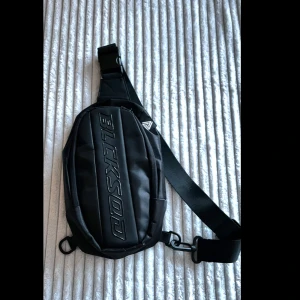 Svart axelväska från BLCKSOQ - Snygg svart axelväska från BLCKSOQ med justerbart band och dragkedja. Väskan har en stilren design med broderad logga framtill och flera fack för dina grejer. Perfekt för att bära mobilen, plånboken och andra småprylar när du är på språng.