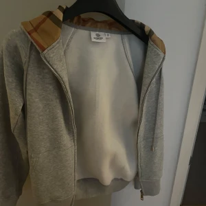 Grå Burberry hoodie med dragkedja - Snygg grå hoodie från Burberry med klassiskt rutigt foder i huvan och broderad logga på bröstet. Tröjan har dragkedja framtill, fickor och ribbade muddar. Perfekt för dig som gillar stilrena och exklusiva plagg med en twist.