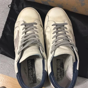 Vita sneakers från Golden Goose - Snygga vita sneakers från Golden Goose med grå snörning och ikonisk stjärndetalj i mocka på sidan. Skorna har en klassisk låg profil, rund tå och mörkblå häl. Perfekta för en avslappnad och trendig stil.