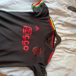 Ajax Reggae Bob Marley fotbollströja Adidas - Ajax svart fotbollströja från Adidas med röd Ziggo-logga och klubbmärke framtill. Färgglada ränder i rött, gult och grönt på ärmar och axlar. Texten 'One Love One Heart One Destiny' på ryggen. Tillverkad i lätt och ventilerande material.