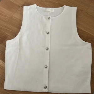 Vit väst med knappar från H&M - Helt ny vit väst från H&M i enkel och stilren design. Västen är ärmlös, har rund halsringning och stängs med fem silvriga knappar framtill. Materialet är mjukt och lätt, perfekt för att bygga lager eller bära som den är.