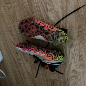 Fotbollsskor Nike Hypervenom - Säljer ett par Nike Hypervenom fotbollsskor med högt skaft och snörning. Skorna är vita med svarta, orange och neongula detaljer samt svart Nike-logga. Sulan har ett coolt mönster i orange, rosa och gult. Perfekta för dig som vill sticka ut på planen. Storlek 40