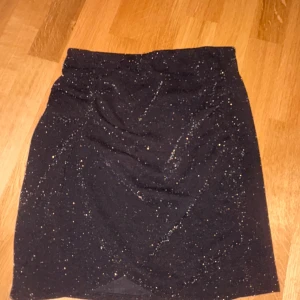 Svart glittrig kjol från Kappahl XS - Svart kortkjol från Kappahl i storlek XS med glittriga detaljer och lätt omlott-effekt framtill. Kjolen har en snygg drapering och är perfekt för dig som vill sticka ut lite extra. Materialet känns mjukt och stretchigt.