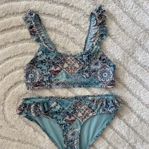 Blåmönstrad bikini från KappAhl - Endast använd en gång, så nyskick! Superfin bikini från KappAhl i storlek 158/164 med blått och turkost patchworkmönster. Både överdel och underdel har volangdetaljer på axelband och sidor. Materialet är mjukt och stretchigt, perfekt för sommardagar vid stranden eller poolen.