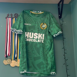 Hammarby grön matchtröja Craft - Säljer en grön Hammarby fotbollströja från Craft med vit logga och tryck 'HUSKI CHOCOLATE' på bröstet. Tröjan har korta ärmar, unikt geometriskt mönster i olika gröna nyanser och detaljer i vitt vid ärmslut och hals. Materialet är lätt och andas, perfekt för match eller träning.
