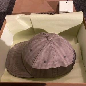 Beige keps från Burberry - Snygg beige keps från Burberry med diskret rutmönster och broderad logga baktill. Klassisk böjd skärm och justerbar rem bak. Tillverkad i bomull för en bekväm och stilren look. Perfekt accessoar för att lyfta din outfit.