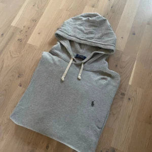Grå hoodie från Polo Ralph Lauren - Snygg grå hoodie från Polo Ralph Lauren med klassisk huva och snörning. Tröjan har en enkel design med den ikoniska lilla loggan broderad på bröstet. Perfekt för en chill och stilren look. Tillverkad i mjuk bomull för extra komfort.