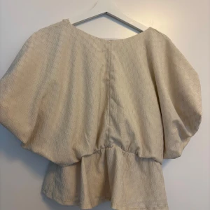 Beige peplum topp med v-ringning från NA-KD - Säljer en beige peplum topp från NA-KD med v-ringning och korta, vida ärmar. Toppen har en lätt struktur i tyget och markerad midja som ger en snygg siluett. Perfekt att styla med både jeans och kostymbyxor för en trendig look.