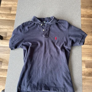 Mörkblå pikétröja från Polo Ralph Lauren - Klassisk mörkblå pikétröja från Polo Ralph Lauren med röd broderad logga på bröstet. Tröjan har korta ärmar, krage och knappar framtill. Tillverkad i mjuk bomull som är skön mot huden. Perfekt för en avslappnad men ändå stilren look.
