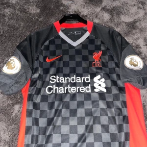 Liverpool bortatröja Virgil 4 Nike L - Liverpool FC bortatröja med Virgil van Dijk #4, Nike Dri-FIT. Svart och grått rutmönster, röda detaljer på krage och sidor, Premier League-märken på ärmarna och klubbmärke på bröstet. Tillverkad i funktionsmaterial som andas.