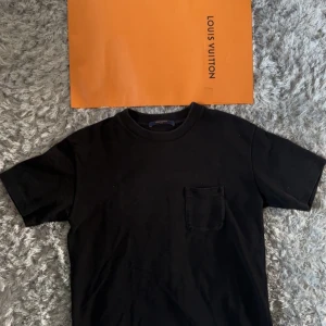 Svart t-shirt från Louis Vuitton - Svart t-shirt från Louis Vuitton med klassisk passform, rund halsringning och en liten bröstficka. T-shirten har diskret Louis Vuitton-detalj i metall vid fållen och är tillverkad i mjuk bomull. Perfekt för dig som gillar stilrena och exklusiva plagg.