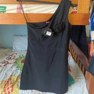 Svart one shoulder klänning med ros - Snygg svart kortklänning med one shoulder-design och dekorativ tygros på axeln. Klänningen har en stilren och figurnära passform, perfekt för dig som vill sticka ut. Enkel men elegant look som passar till många tillfällen.