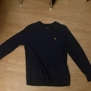 Mörkblå tröja från Lyle & Scott - Snygg mörkblå tröja från Lyle & Scott i 100% bomull. Klassisk modell med rund hals och lång ärm, prydd med den ikoniska gula loggan på bröstet. Perfekt för dig som gillar stilren och enkel design och är mycket bra.