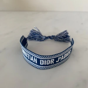 Blått Christian Dior J'Adior armband - Snyggt blått armband från Christian Dior med texten 'J'Adior' broderad i vitt. Armbandet har justerbar knytning med tofsar i ändarna och är gjort i ett mjukt tygmaterial. Perfekt accessoar för att lyfta din outfit med en lyxig touch.