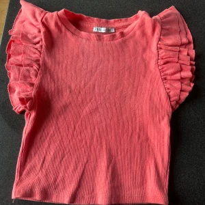 Rosa volangtopp från Zara - Supersöt rosa topp från Zara med ribbad struktur och volangdetaljer på axlarna. Kortärmad modell i mjuk bomull som ger en skön känsla och snygg passform. Perfekt för dig som vill ha en färgglad och trendig look.