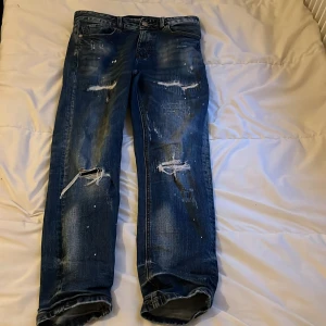 Blå distressed jeans Supply & Demand XS - Säljer ett par blå jeans från Supply & Demand i storlek XS. Jeansen har slitna detaljer, hål på knäna och coola färgstänk. Klassisk femficksmodell med normal passform och uppvikta benslut. Perfekta för en streetstyle-look. De är nästan helt oanvända jag har bara använt dem några gånger 