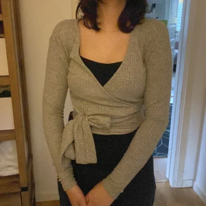 Grå ribbad omlottkofta från Zara - Supersnygg grå ribbad omlottkofta från Zara med lång ärm och knyt i midjan. Koftan har en croppad passform och djup v-ringning, perfekt att styla över ett linne eller klänning. Mjuk och stretchig, passar året runt.