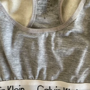 Grå linnetopp från Calvin Klein - Sportig grå topp från Calvin Klein med klassisk logotyp på det vita resårbandet nedtill. Toppen har brottarrygg och är tillverkad i mjuk bomullsblandning som sitter skönt mot kroppen. Perfekt för en chill look eller till träning.