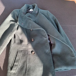 Mörkgrön skepparkappa med hög krage - Stilren mörkgrön och figursydd skepparkappa från H&M med dubbelknäppning och hög krage som kan bäras uppfälld. Jackan har snygga knappar framtill, raka fickor och är tillverkad i ett mjukt ullblandat tyg. Perfekt för uppklädda tillfällen eller en stilren vardag. Den är i storlek 38 men passar mig som i vanliga fall har M