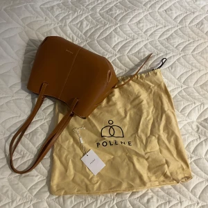 Polène cyme bag camel - Säljer en snygg handväska från Polène i brunt skinn, camel med korta och långa handtag. Väskan har magnetlås, innerficka med dragkedja och kommer med original dustbag, liten pouch och lapp. Materialet är i perfekt skick, handtagen har smått slitage.