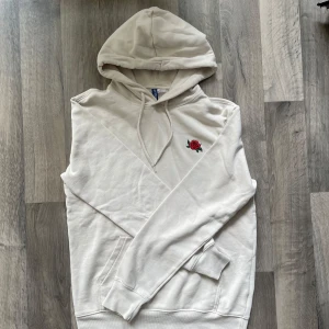 Beige hoodie från H&M - Snygg beige hoodie från H&M med en röd ros på bröstet. Perfekt för chill dagar och enkel att matcha med jeans eller joggers.