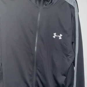 Svart vindjacka från Under Armour - Grå vindjacka från Under Armour med gråa detaljer längs axlarna och ärmarna. Jackan har dragkedja framtill, hög krage och det klassiska Under Armour-loggan på bröstet. Tillverkad i lätt polyester, perfekt för träning eller chill dagar. Aldrig använd 