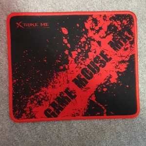 Mous pad - Bilderna visar en gaming-mousepad från Xtrike Me. Produkten har en svart och röd design med texten 'GAME MOUSE PAD' och är perfekt för gaming-setupen. Den har sydda kanter för extra hållbarhet och en halkfri undersida.